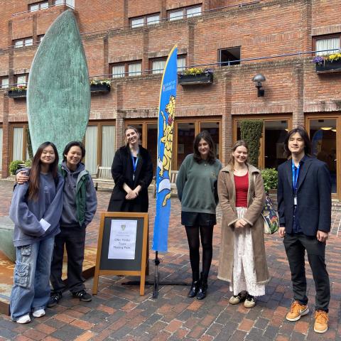 A group of student ambassadors in Front Court next to a Robinson crest flag and a sign which reads 'meeting point for offer holder tours'
